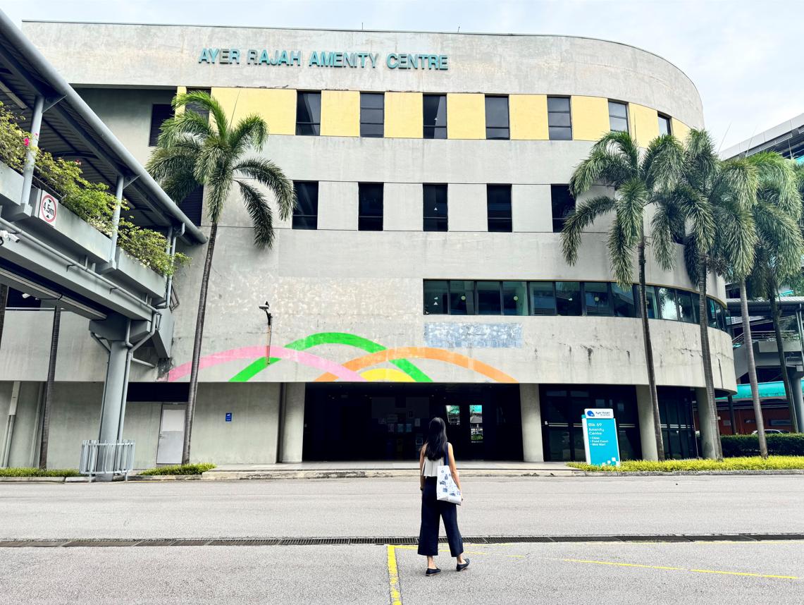 Ayer Rajah Amenity Centre with a person crossing the road in front of it, with palm trees on each side.